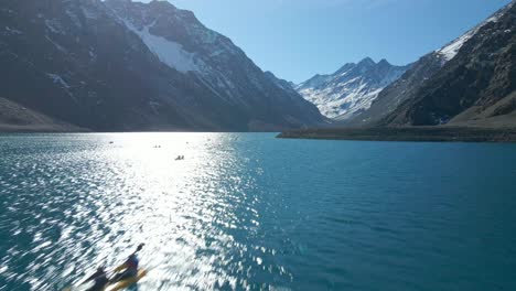 Vista-Aérea-De-Personas-En-Kayaks-En-La-Laguna-Del-Inca,-En-Los-Andes-Chilenos,-Montañas-Nevadas-En-Un-Día-Soleado-En-El-Fondo