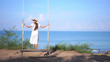 A-young-woman-in-a-billowy-sundress-stands-on-a-huge-wooden-rope-swing-looking-out-on-the-ocean-as-the-waves-crash-on-the-shore