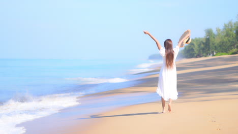 A-happy-young-woman,-in-a-white-flowing-sundress-carrying-a-large-sun-hat,-dances-along-the-beach-as-the-waves-roll-in