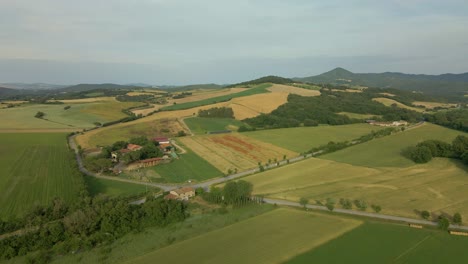 Imágenes-Aéreas-De-Toscana-En-Italia-Campos-Cultivados-Verano,-Pequeña-Granja-Con-Montañas-Al-Fondo-Coloridos-Campos-Sembrados