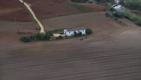 Vista-Aérea-Hacia-El-Campo-Rural-Con-Casa-De-Campo-Y-Gente-Montando-A-Caballo