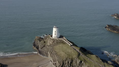 An-aerial-view-of-Twr-Mawr-Lighthouse-on-Ynys-Llanddwyn-island,-flying-left-to-right-around-the-lighthouse-while-elevating-and-zooming-out,-Anglesey,-North-Wales,-UK
