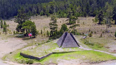 Aerial-orbit-shot-of-Colossal-Pyramid,-Valle-Nuevo-and-waving-flag-surrounded-by-forest-in-Dominican-Republic