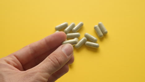 Left-Male-Hand-Holding-Lions-Mane-Supplement-Pill-In-Between-Fingers-With-Yellow-Background-With-Pills-On-Table