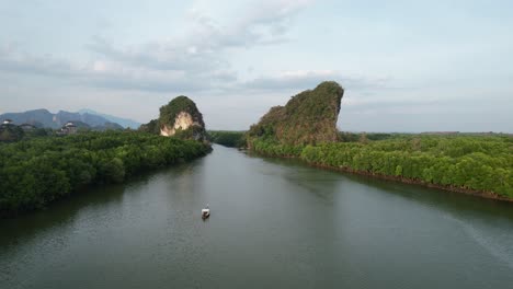 aerial-drone-flying-backwards-revealing-a-Thai-longtail-boat-on-a-river-between-a-two-large-green-limestone-mountain-mountains-and-mangrove-forest-in-Krabi-Town-Thailand-at-sunset