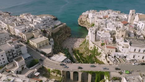 Slow-aerial-flyover-of-car-traffic-going-over-a-bridge-in-Polignano-a-Mare,-Italy-with-a-view-of-the-sea-and-beach