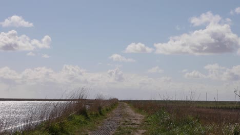 Footpath-water-on-the-left-marsh-on-the-right,-horizon-and-skyline-with-fluffy-clouds-and-soft-blue-sky