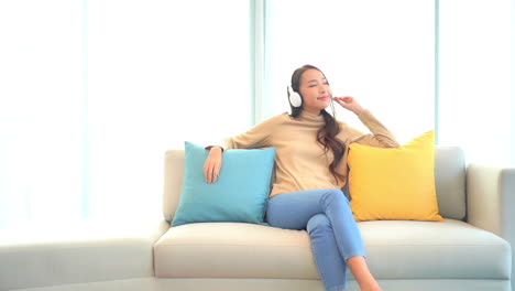 A-pretty-young-woman-sitting-on-a-couch-between-colorful-decorative-pillows-while-listening-to-music-through-her-headphones