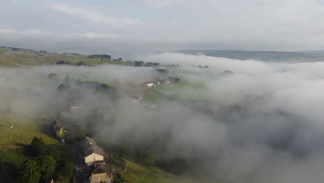 Establishing-shot,-Foggy-morning-over-the-Yorkshire-Dales,-meadow-near-a-rural-village,-aerial-view-landscape-moving-through-the-clouds-when-it-is-sunny