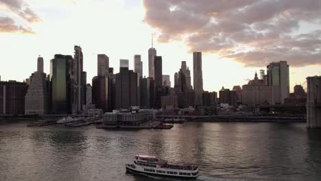 Aerial-view-over-a-ferry-on-East-river-in-front-of-the-Lower-Manhattan-skyline,-sunset-in-NYC,-USA---rising,-drone-shot