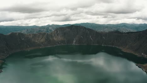 Aerial-backwards-shot-of-natural-crater-lake-surrounded-by-volcano-rocks-during-cloudy-day