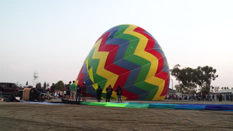 Der-Heißluftballon-Liegt-Auf-Der-Seite,-Während-Er-Befüllt-Wird-Und-Sich-Auf-Den-Start-Vorbereitet,-Während-Eine-Menschenmenge-In-Der-Nähe-Steht-Und-Erwartungsvoll-Zuschaut