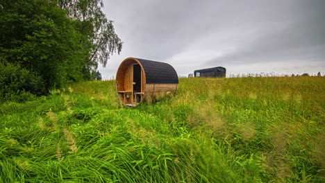 Static-shot-of-rectangular-cabin-and-barrel-sauna-with-green-grass-been-cut-by-grass-cutter-machine-during-spring-time-in-timelapse