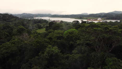 Aerials-footage-of-a-cargo-ship-sailing-down-a-vast-river-through-a-lush-forest-passing-by-a-quaint-harbor