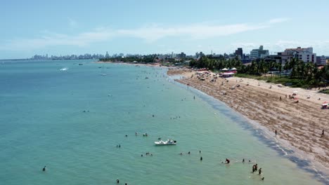 Tilt-up-aerial-shot-of-Bessa-beach-during-low-tide-in-the-tropical-capital-city-of-Joao-Pessoa,-Paraiba,-Brazil-with-people-enjoying-the-ocean-and-fishing-boats-at-shore-on-a-summer-day