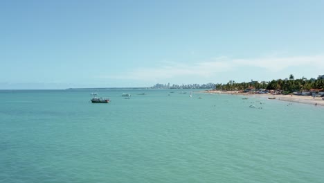 Trucking-left-aerial-wide-shot-of-the-tropical-Bessa-beach-in-the-coastal-capital-city-of-Joao-Pessoa,-Paraiba,-Brazil-with-people-enjoying-the-ocean-and-fishing-boats-at-shore-on-a-sunny-summer-day