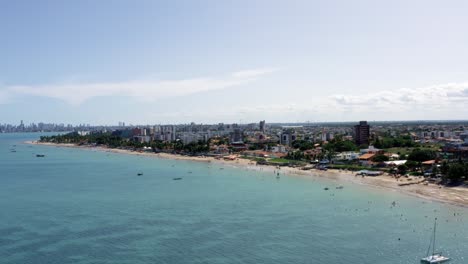 Rising-trucking-right-aerial-extreme-wide-shot-of-the-tropical-Bessa-beach-in-the-capital-city-of-Joao-Pessoa,-Paraiba,-Brazil-with-people-enjoying-the-ocean-surrounded-by-palm-trees-and-buildings
