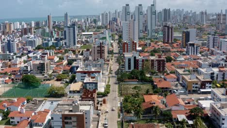 Tilt-up-rising-aerial-drone-shot-of-the-tropical-beach-town-of-Cabedelo-with-the-capital-city-of-Joao-Pessoa,-Paraiba,-Brazil-in-the-distance-with-cityscape-along-the-coastline-and-rural-homes-below