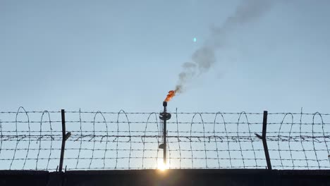 Industrial-Gas-flare-behind-wired-fence,-zoom-out,-day