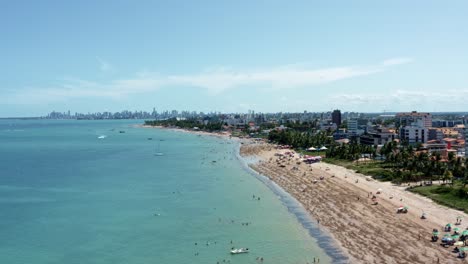 Descending-aerial-shot-of-Bessa-beach-during-low-tide-in-the-tropical-capital-city-of-Joao-Pessoa,-Paraiba,-Brazil-with-people-enjoying-the-ocean-and-fishing-boats-at-shore-on-a-summer-day