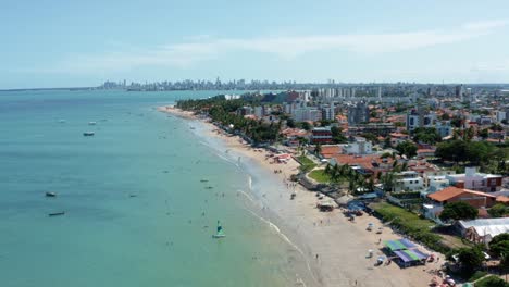Dolly-in-tilting-up-aerial-extreme-wide-shot-of-the-tropical-Bessa-beach-in-the-capital-city-of-Joao-Pessoa,-Paraiba,-Brazil-with-people-enjoying-the-ocean-surrounded-by-palm-trees-and-buildings