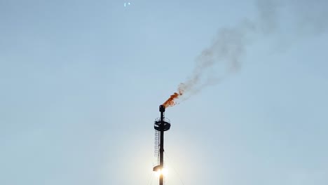 Close-up-of-Gas-flare-top-with-flame-burning-and-smokestack-in-front-of-sun