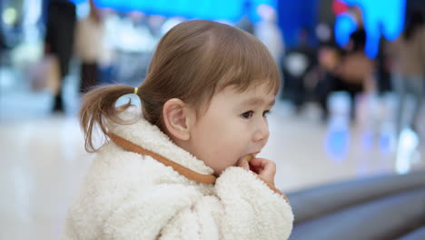 Side-View-Of-A-3-Year-Old-Toddler-Girl-Sitting-Eating-Bread-Inside-A-Shopping-Mall