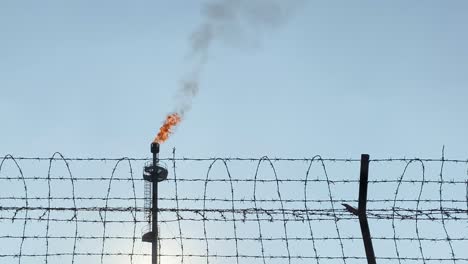A-gas-plant-chimney-burns-behind-barbed-wire,-representing-the-economic-war-over-fossil-fuel-prices