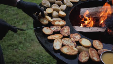 Male-chef-preparing-potatoes-and-vegetables-for-quests-on-open-fire-grill-in-the-backyard-on-summer-evening-with-black-gloves-on-close-up