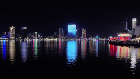 Da-Nang-City-Downtown-by-Night,-Vietnam,-Timelapse-Panoramic-View-of-Han-River,-illuminated-Buildings-and-Towers,-Boats-Sailing-Passing,-Tourists-on-Love-Pier-Famous-Tourist-Destination