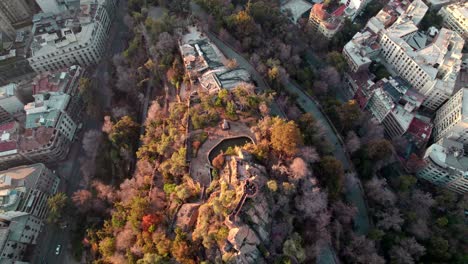 Aerial-rising-over-top-of-Santa-Lucia-Hill-covered-with-autumnal-trees-and-Pedro-de-Valdivia-Square-between-Santiago-buildings,-Chile