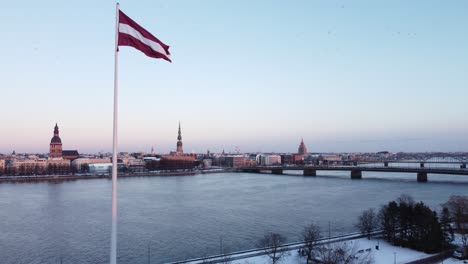 Scenic-aerial-view-of-Latvian-Flag-on-pole,-flying-forward-reveal-cityscape