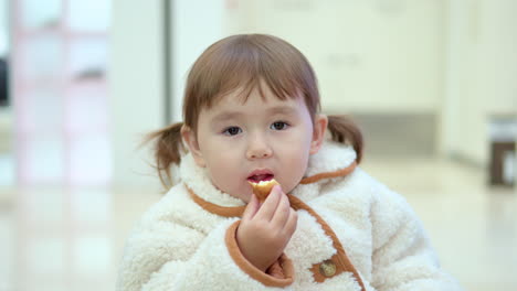 Cute-3-Years-Old-Toddler-In-Ponytails-And-Fluffy-Coat-Enjoys-Eating-Bread-Inside-A-Shopping-Mall-With-Blurred-View-Of-People-Walking-Behind