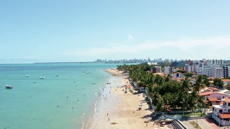 Rising-aerial-wide-shot-of-Bessa-beach-in-the-tropical-capital-Joao-Pessoa,-Brazil-in-the-state-of-Paraiba-with-Brazilians-and-tourists-enjoying-the-ocean-and-fishing-boats-at-shore-on-a-summer-day