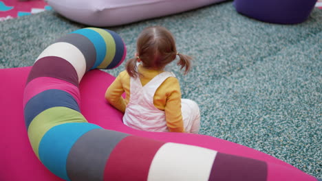 Ein-3-jähriges-Mädchen-Mit-Blonden-Haaren-Und-Pferdeschwänzen-Sitzt-Auf-Einer-Zartrosa-Couch-Mit-Einem-Bunten-Langen-Kissen-Im-Spielbereich-In-Einem-Einkaufszentrum