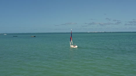 Rotierende-Luftaufnahme,-Die-Einem-Farbenfrohen-Segelboot-In-Der-Nähe-Des-Tropischen-Strandes-Bessa-In-Der-Küstenhauptstadt-Joao-Pessoa,-Paraiba,-Brasilien,-Folgt,-Mit-Der-Berühmten-Roten-Sandinsel-Im-Hintergrund