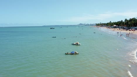 Dolly-in-aerial-wide-shot-of-the-tropical-Bessa-beach-in-the-coastal-capital-city-of-Joao-Pessoa,-Paraiba,-Brazil-with-people-enjoying-the-ocean-and-fishing-boats-at-shore-on-a-warm-sunny-summer-day