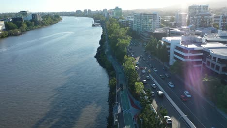 Aerial-view-of-Brisbane's-beautiful-river-as-you-fly-along-with-this-panning-drone-shot,-capturing-Coronation-Drive-and-bike-paths-with-busy-traffic,-featuring-a-stunning-afternoon-lens-flare