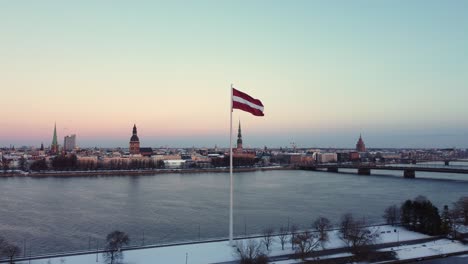 Latvian-flag-waving-by-Daugava-river-with-cityscape,-winter-snowy-city,-aerial