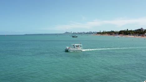 Trucking-left-aerial-shot-flying-past-a-tourist-boat-taking-people-to-the-famous-Red-Sand-Island-from-Bessa-beach-in-the-tropical-capital-city-of-Joao-Pessoa,-Paraiba,-Brazil-on-a-sunny-summer-day