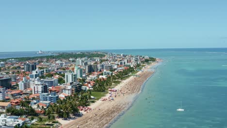 Trucking-right-aerial-shot-of-Bessa-beach-in-the-tropical-capital-Joao-Pessoa,-Brazil-in-the-state-of-Paraiba-with-locals-and-tourists-enjoying-the-ocean-and-fishing-boats-at-shore-on-a-summer-day