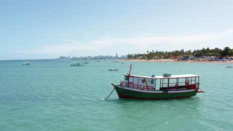 Luftdrohnenaufnahme-Eines-Kleinen-Ausflugsbootes,-Das-In-Der-Nähe-Des-Tropischen-Bessa-Strandes-Anlegt,-Mit-Wolkenkratzern-Aus-Der-Küstenhauptstadt-Joao-Pessoa,-Paraiba,-Brasilien-Im-Hintergrund-An-Einem-Sonnigen-Sommertag