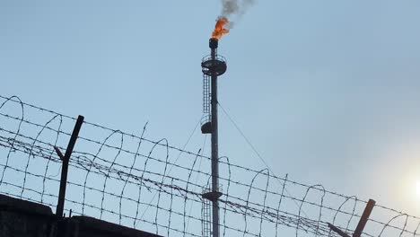 Low-angle-view-of-Flare-stack-refinery-behind-wired-fence,-tilt-up-reveals-burning-flame-and-smokstack