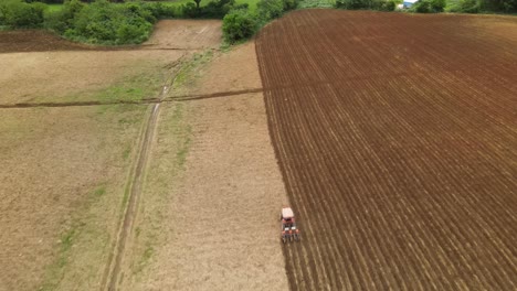 An-aerial-footage-following-an-orange-tractor-tilling-a-farmland-also-revealing-a-treeline,-some-birds,-Muak-Klek,-Thailand