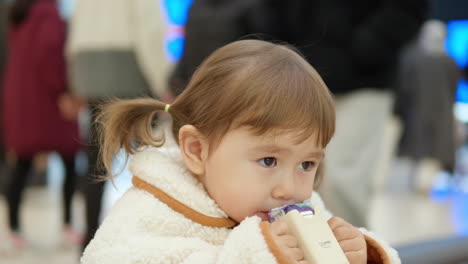 3-year-old-Female-Child-Drinking-Juice-In-The-Shopping-Mall-Cafe,-Shoppers-Walking-By-In-Blurry-Background