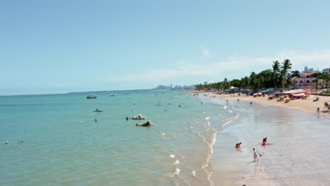 Dolly-in-aerial-shot-of-Bessa-beach-in-the-tropical-capital-Joao-Pessoa,-Brazil-in-the-state-of-Paraiba-with-Brazilians-and-tourists-enjoying-the-ocean-and-fishing-boats-at-shore-on-a-summer-day