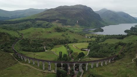 Aerial:-Train-approaches-iconic-curving-Glenfinnan-Viaduct,-Scotland