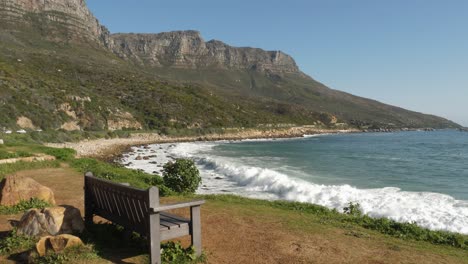 One-lone-bench-overlooks-rocky-beach-and-mountains-in-Cape-town,-RSA