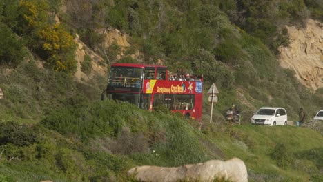 Red-doubledecker-bus-tours-along-mountainous-beach-road,-Cape-town