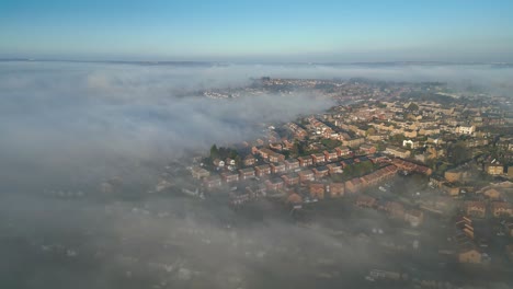 Aerial-drone-point-of-view-UK-cityscape-during-sunrise,-early-morning-landscape,-residential-building-rooftops-at-dense-fog,-misty-weather